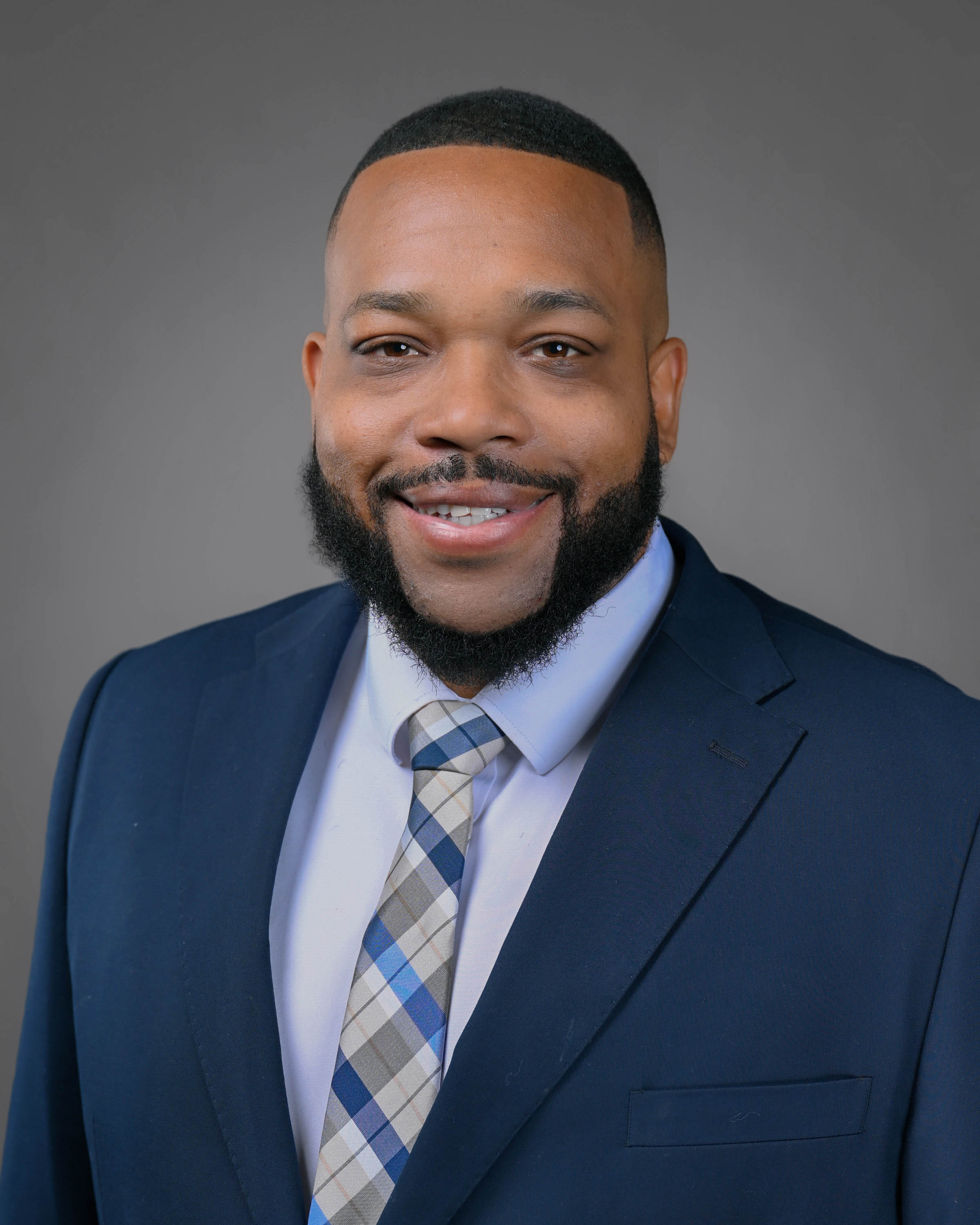 Man smiling with short black hair and a beard wearing a blue jacket and tie